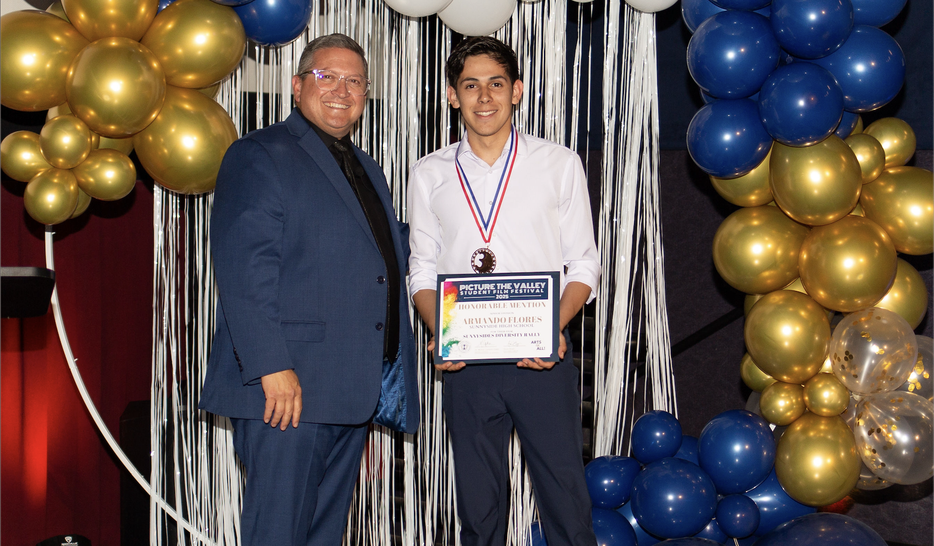 Young man standing in front of a blue, gold, and white balloon arch holding an award certificate and wearing a medallion.